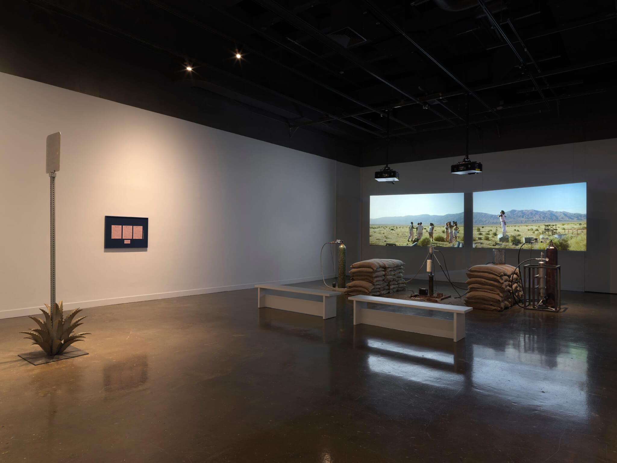 Inside a dimly lit gallery space, two projections sit behind a rocket test installation containing sandbags and canisters. In the foreground to the left is a metal sign inside an agave plant and a blue frame with pink paper mounted.
